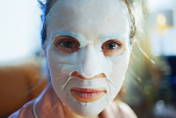 Portrait de femme élégante de 40 ans en pyjama avec masque facial en feuille blanche sur le visage à la maison moderne dans la journée ensoleillée d'hiver
. - Photo, image