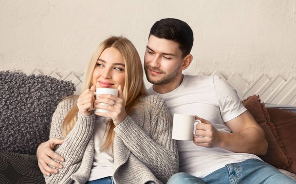 Affectionate couple with hot drinks cuddling on sofa in living room - Photo, Image