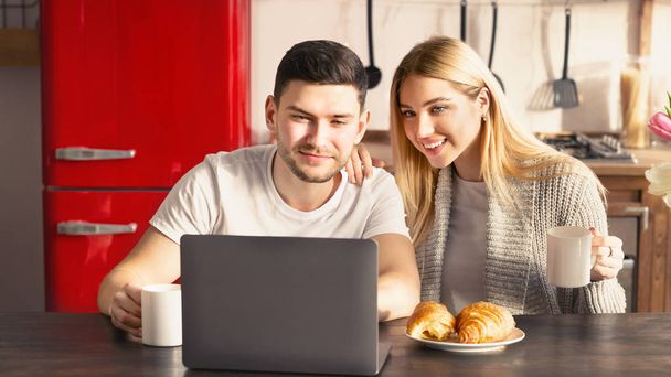 Beautiful young couple with breakfast and laptop at table in kitchen - Zdjęcie, obraz