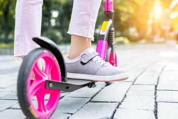 Bottom part of young adult female person wearing pink casual jeans and sneakers riding push kick scooter at city street on summer day outdoor. Teenager girl having fun enjoy sport activities outside. - Photo, image