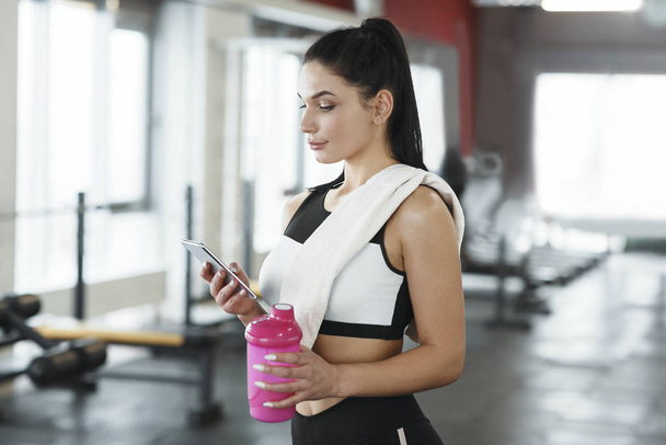 Молода жінка зі смартфоном і пляшкою води або білковим струмом у спортзалі
 - Фото, зображення