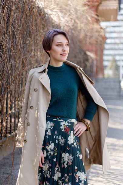 Portrait shooting of a attractive stylish short cut woman, Girl walks in the city outdoors. Stylish modern and feminine image, style. Girl in a beige cloak or coat and a green dress - Фото, изображение