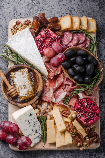 Traditional italian antipasto plate. Assorted cheeses on wooden cutting board. Brie cheese, cheddar slices, gogonzola, walnuts grapes, olives, prosciutto, rosemary and glass of red wine. top view - Photo, Image