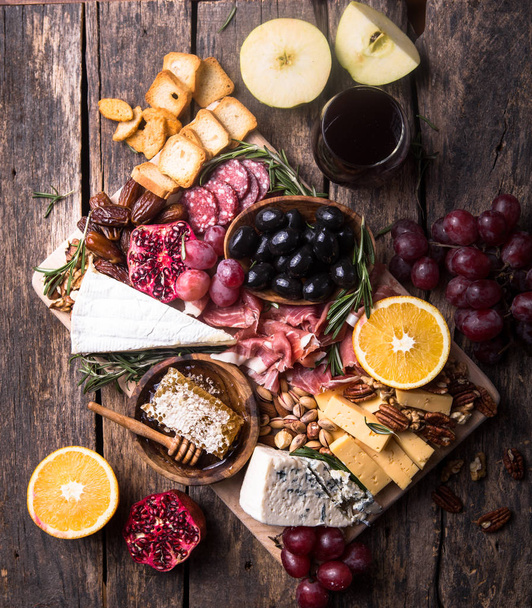 Traditional italian antipasto plate. Assorted cheeses on wooden cutting board. Brie cheese, cheddar slices, gogonzola, walnuts grapes, olives, prosciutto, rosemary and glass of red wine. top view - Foto, Bild