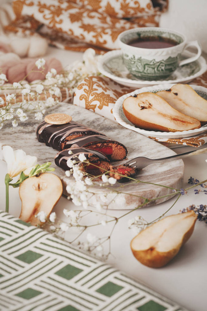 Chocolate coated dessert on a marble stand with fork.The table are decorated cut pears, small white flowers and different napkins with patterns.Table setting for breakfast. Vintage dessert concept. - Photo, Image