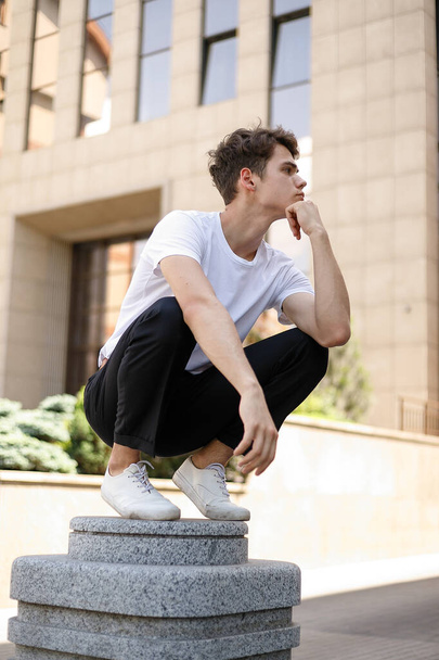 Elegant young man in a fashionable black shirt in a white stylish t-shirt in black pants with a trendy hairstyle rests near a modern business center. Attractive guy in the street on a summer day. - Zdjęcie, obraz