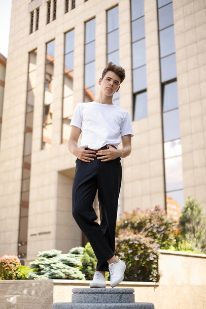 Elegant young man in a fashionable black shirt in a white stylish t-shirt in black pants with a trendy hairstyle rests near a modern business center. Attractive guy in the street on a summer day. - Foto, Bild