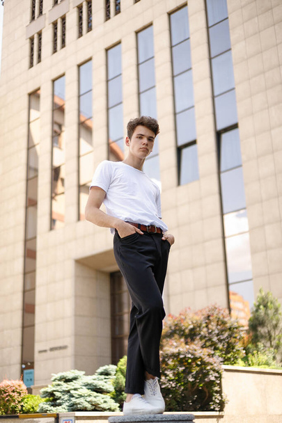Elegant young man in a fashionable black shirt in a white stylish t-shirt in black pants with a trendy hairstyle rests near a modern business center. Attractive guy in the street on a summer day. - Photo, image