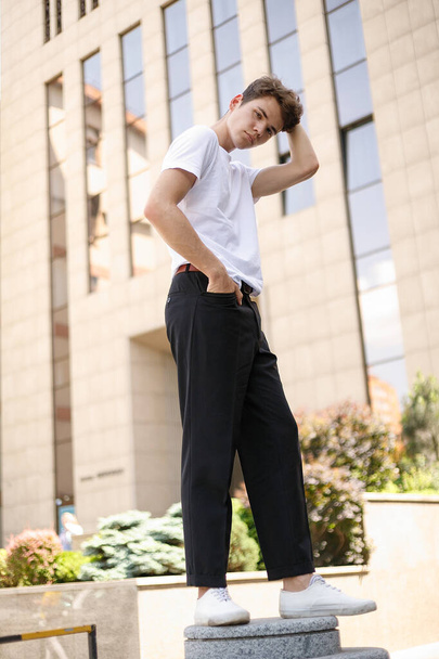 Eleganter junger Mann in modischem schwarzen Hemd, weißem stylischem T-Shirt, schwarzer Hose und trendiger Frisur ruht in der Nähe eines modernen Businesscenters. Attraktiver Kerl auf der Straße an einem Sommertag. - Foto, Bild