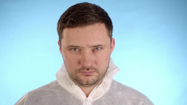 Kaukasischer bärtiger Mann in weißem Schutzanzug vor blauem Hintergrund blickt nach unten und dann in die Kamera - Filmmaterial, Video