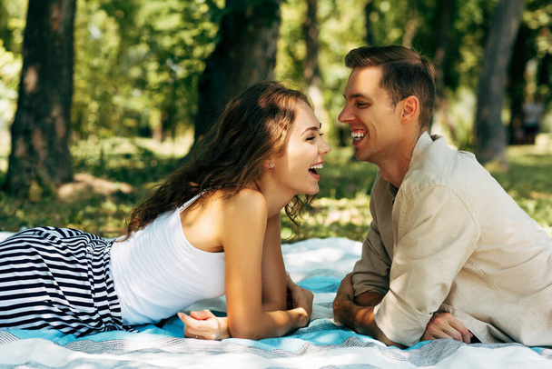 Side view image of happy couple in love laughing and dating outdoors at the park on a sunny day. Young man ansd woman lying on the blanket on the grass during picnic in the nature. Happy relationship - Photo, Image