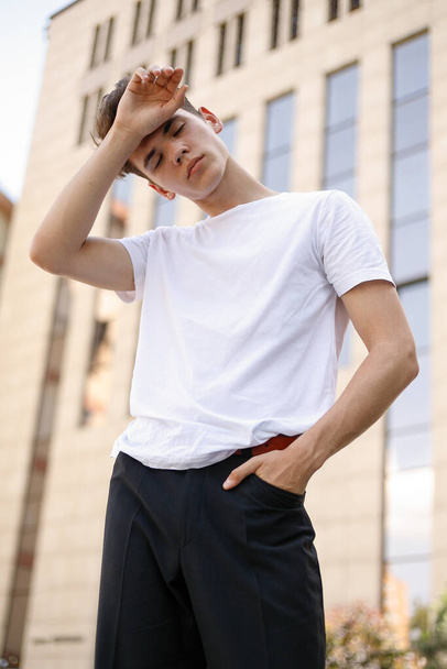 Elegant young man in a fashionable black shirt in a white stylish t-shirt in black pants with a trendy hairstyle rests near a modern business center. Attractive guy in the street on a summer day. - Foto, immagini