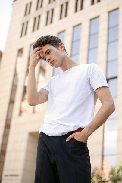Elegant young man in a fashionable black shirt in a white stylish t-shirt in black pants with a trendy hairstyle rests near a modern business center. Attractive guy in the street on a summer day. - Fotoğraf, Görsel