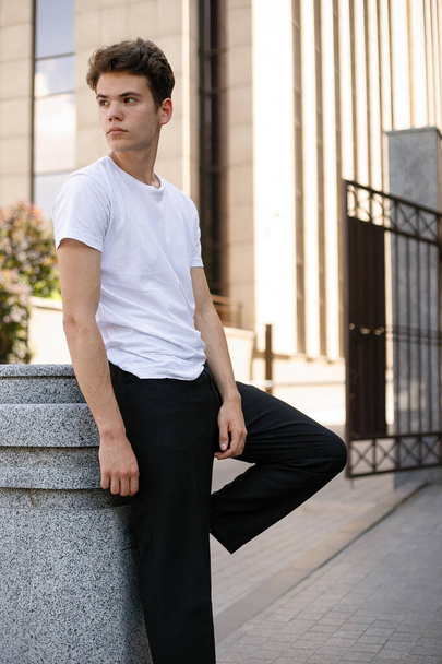 Elegant young man in a fashionable black shirt in a white stylish t-shirt in black pants with a trendy hairstyle rests near a modern business center. Attractive guy in the street on a summer day. - Foto, afbeelding