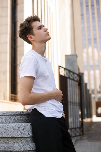 Elegant young man in a fashionable black shirt in a white stylish t-shirt in black pants with a trendy hairstyle rests near a modern business center. Attractive guy in the street on a summer day. - Fotoğraf, Görsel