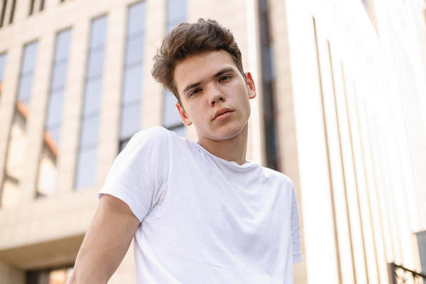 Elegant young man smile in a fashionable black shirt in a white stylish t-shirt in black pants with a trendy hairstyle rests near a modern business center. close-up portrait - Foto, Bild