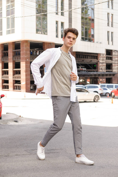 guy model with a stylish haircut posing outdoors in a white shirt and gray trousers. trendy hairstyle rests near a modern business center. Attractive guy in the street on a summer day. - Фото, изображение