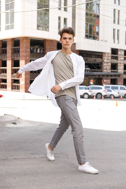 guy model with a stylish haircut posing outdoors in a white shirt and gray trousers. trendy hairstyle rests near a modern business center. Attractive guy in the street on a summer day. - Photo, image