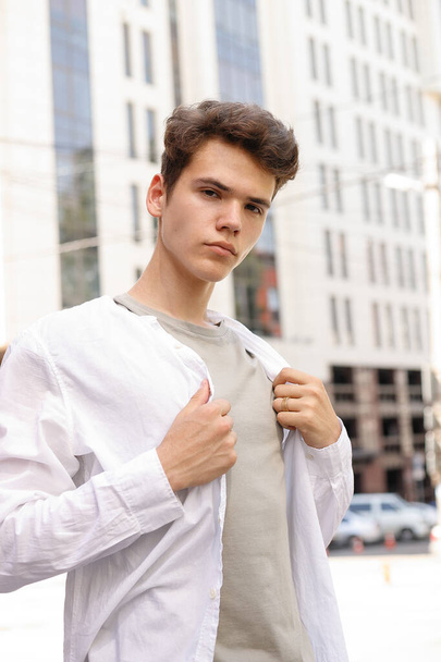 guy model with a stylish haircut posing outdoors in a white shirt and gray trousers. trendy hairstyle rests near a modern business center. Attractive guy in the street on a summer day. - Foto, afbeelding