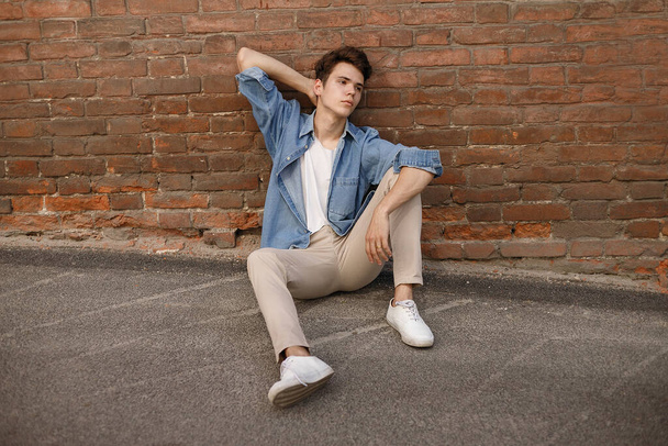 Young hipster sits in his blue denim button up shirt showing an unlabeled white cotton t-shirt against a brick wall background - Фото, зображення