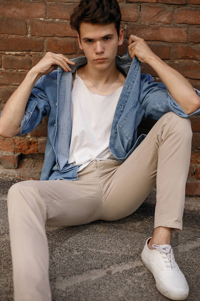 Young hipster sits in his blue denim button up shirt showing an unlabeled white cotton t-shirt against a brick wall background - Fotó, kép