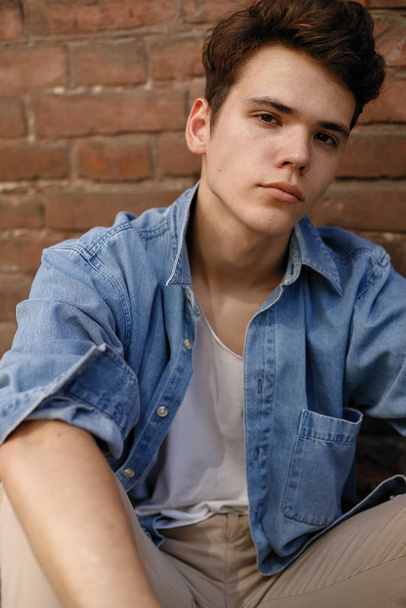 Young hipster sits in his blue denim button up shirt showing an unlabeled white cotton t-shirt against a brick wall background - Foto, imagen