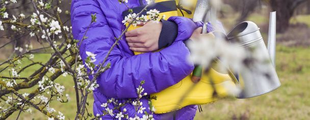 The girl holds bright yellow rubber boots, a watering can, a spade in her hands. The flowering period of trees in the gardens and working hours on personal plots. Spring. Branches out of focus. Banner - Fotoğraf, Görsel