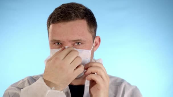 Caucasian bearded man in a white protective jumpsuit on a blue background puts a medical mask on his face, zips his jacket and puts on a hood. - Metraje, vídeo