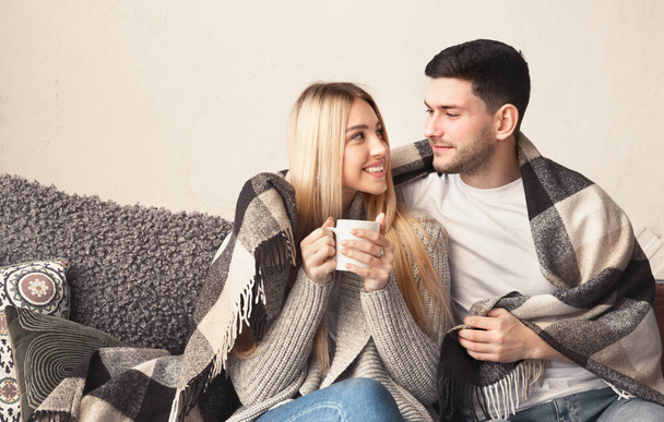 Lächelndes junges Paar mit Heißgetränken kuschelt sich zu Hause unter eine warme Decke, leerer Raum - Foto, Bild