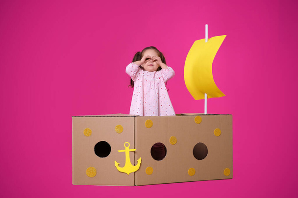 Little child playing with ship made of cardboard box on pink background - Foto, afbeelding
