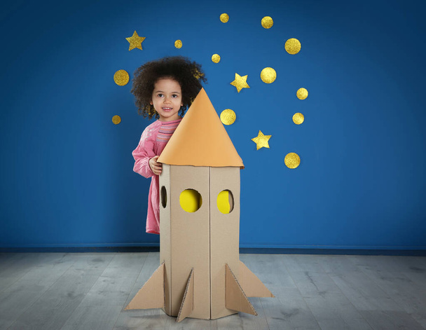 Cute African American child playing with cardboard rocket near blue wall - Foto, immagini