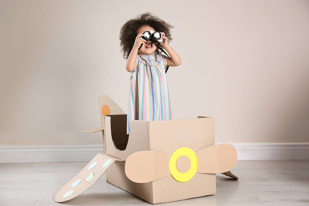 Cute African American child playing with cardboard plane and binoculars near beige wall - Foto, Imagen