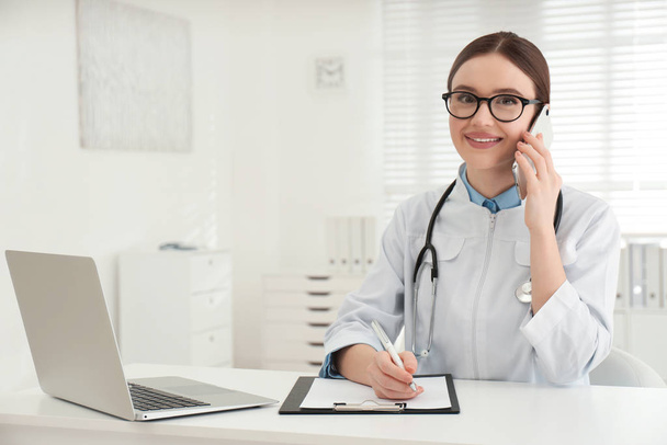 Young female doctor talking on phone at table in office - Foto, immagini