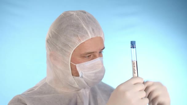 Caucasian man in white protective suit, medical mask, rubber gloves on blue background holds in hands test tube with red liquid and inscription Coronovirus, then shows test tube to camera - Video