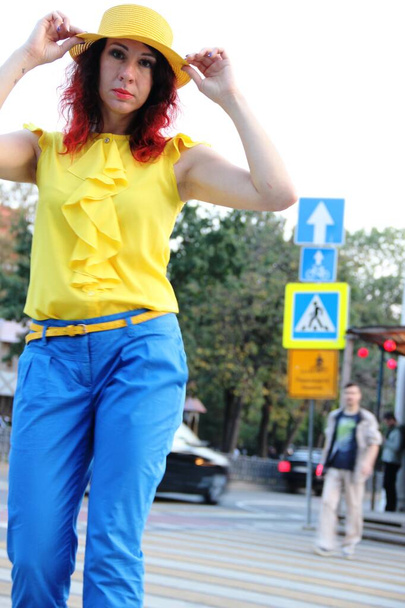 woman with red curly hair, in blue classic trousers, a yellow blouse and a hat in the city, stands on a pedestrian crossing, blur - Foto, afbeelding