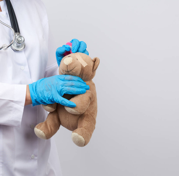 doctor in a white coat and blue latex gloves holds a brown teddy bear without an eye, doctor wraps his head with adhesive plaster - Photo, Image