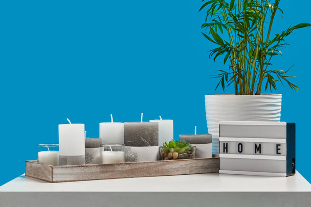 Witte tafel met groene plant en sappig in potten, verschillende grootte kaarsen in houten standaard, lamp met inscriptie huis. Blauwe achtergrond. Sluiten. - Foto, afbeelding