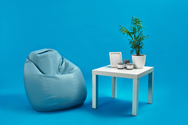 Turquoise armchair bean bag and white coffee table with green flower in a pot, three candles and photo frame on it. Blue studio background. Copy space - Foto, Imagem