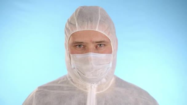 Caucasian man in white protective suit, medical mask, rubber gloves on blue background holds test tube with red liquid and inscription coronovirus, then drops test tube on floor and begins panic - Imágenes, Vídeo