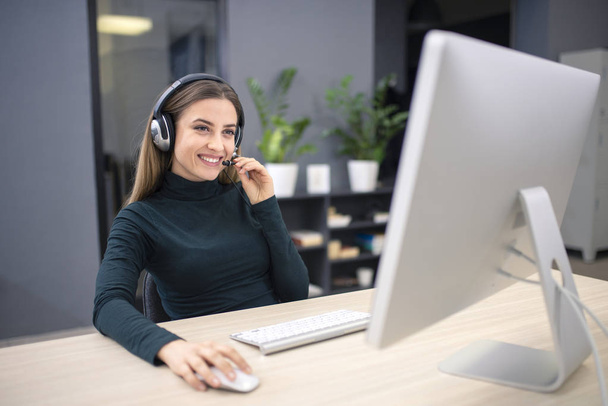 Mladá hezká kancelářská pracovnice s úsměvem při komunikaci se zákazníkem přes internet, nošení sluchátek s mikrofonem - Fotografie, Obrázek
