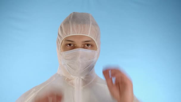 Caucasian bearded man in a white protective jumpsuit on a blue background removes the medical mask from his face and hood, unbuttons the zipper of his jacket and sighs deeply - Séquence, vidéo