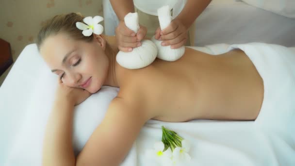 Massagetherapeut gebruik warme kruidenballen massage op de rug voor hulp verlichten van stress en helpt ontspanning. Aantrekkelijk mooi meisje wordt gelukkig, glimlach. Charmante mooie vrouw liggen gevoelig op bed in spa salon. - Video