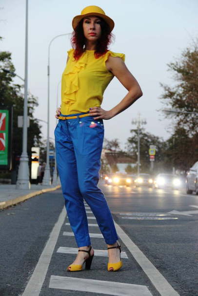 赤い巻き毛の女性、青いクラシックパンツを着て、黄色のブラウスと黄色の帽子は、道路標識に立っている、都市部の夜には、遠くに車のライト、背景がぼやけている - 写真・画像