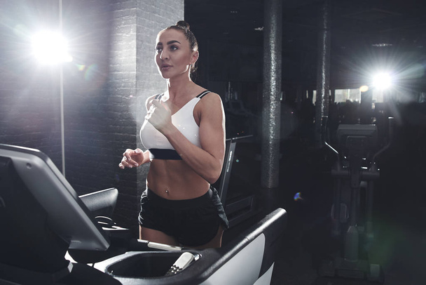 Close portrait fit sexy strong brunette woman in sportswear top and shorts runs on a running track in the gym, cardio workout - 写真・画像