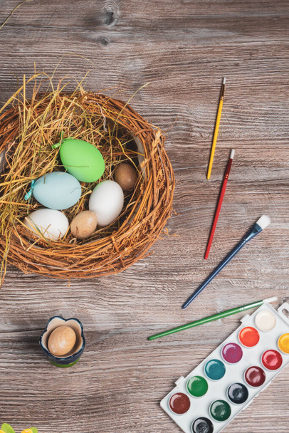 preparing for easter holidays, on wooden table nest with colorful eggs, paints and brushes - Foto, afbeelding