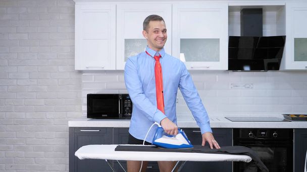 handsome man in tie ironing pants . the kitchen of his house - Фото, изображение