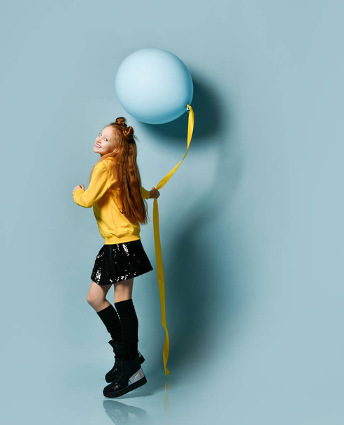 Teenage girl in yellow sweatshirt, black skirt, knee-highs, boots. She holding balloon, posing sideways on blue background. - Photo, Image