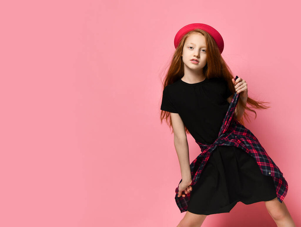 Teenage kid in black dress and red hat. She tightens checkered shirt on her waist while posing against pink background. Close up - Foto, imagen
