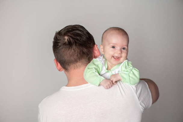 Όμορφος πατέρας κρατώντας ένα χαριτωμένο μικρό χαμογελαστό μωρό στην αγκαλιά του σε ένα γκρι φόντο - Φωτογραφία, εικόνα
