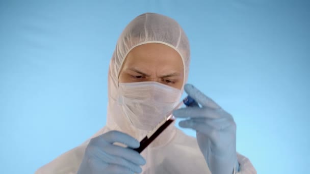 Caucasian man in white protective suit, medical mask, rubber gloves on blue background holds test tube with red liquid and inscription coronovirus, then drops test tube on floor and looks at camera - Footage, Video